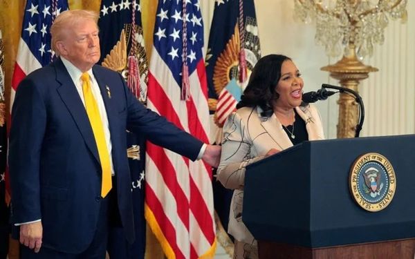 Two people stand on a stage with U.S. flags behind them; a man in a dark suit and yellow tie stands beside a woman speaking at a podium with a presidential seal.