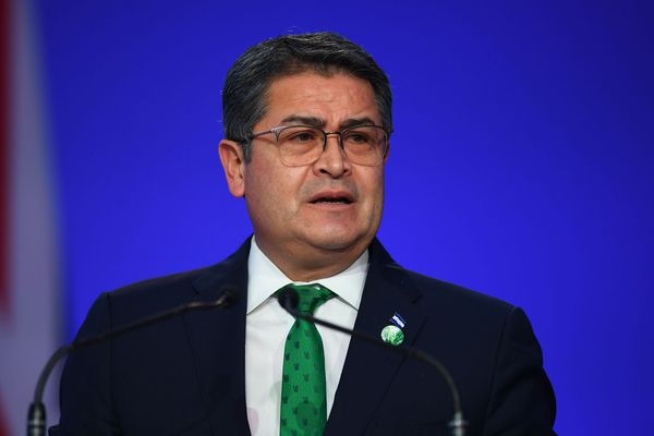 Man in a dark suit and green tie speaks at a podium against a blue backdrop, wearing glasses.