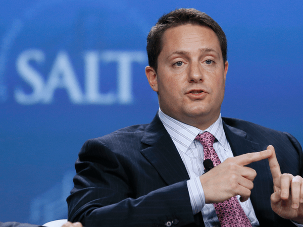 Male conference speaker in a suit, gesturing with his hands on a blue backdrop with a partial 'SALT' logo behind him