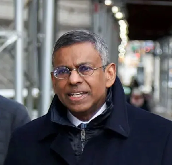 Man wearing round glasses and a dark coat outdoors on a city sidewalk.