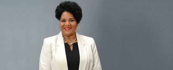 Smiling African American woman in a white blazer and black top, wearing a beaded necklace and hoop earrings, against a grey backdrop.