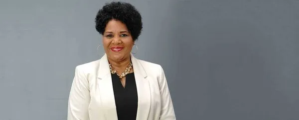 Smiling African American woman in a white blazer and black top, wearing a beaded necklace and hoop earrings, against a grey backdrop.