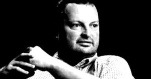 A thoughtful man in a checkered shirt gestures with his hand against a dark background, conveying contemplation and engagement.