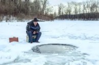 Elderly man sits with a fishing rod at the ice-hole on the lake. Winter fishing