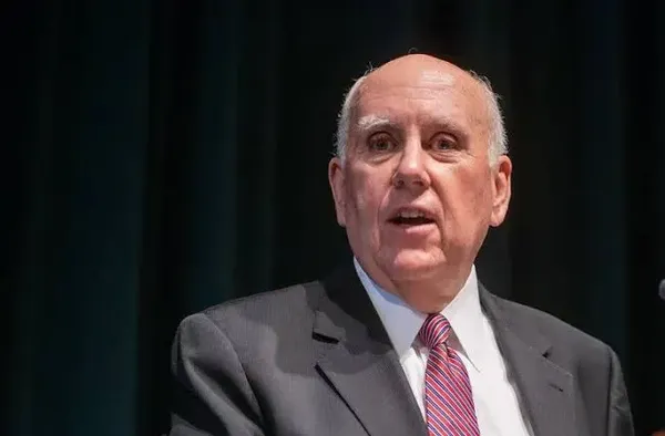 Older man in a suit and striped tie speaks at a podium on a dark stage behind him.