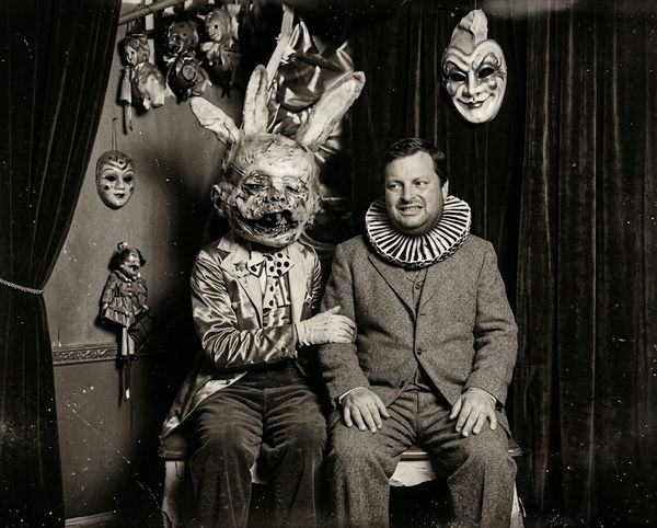 Two men seated on a bench in a theater setting wearing eerie costumes: one with a worn rabbit head mask, the other in a suit with a ruffled clown collar, surrounded by hanging masks and puppets.