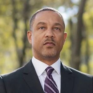 Head-and-shoulders portrait of a man in a dark suit and purple striped tie outdoors, with blurred trees in the background.