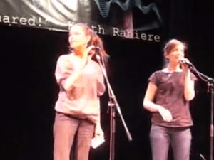 Kristin Kreuk on stage with Nicki Clyne promoting Keith Raniere [see the banner in the background.]