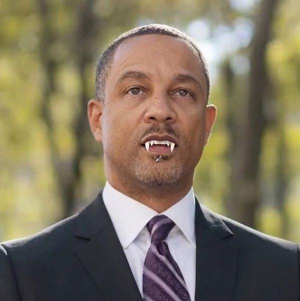 Man in a suit outdoors wearing fake vampire fangs for a costume effect, looking at the camera.